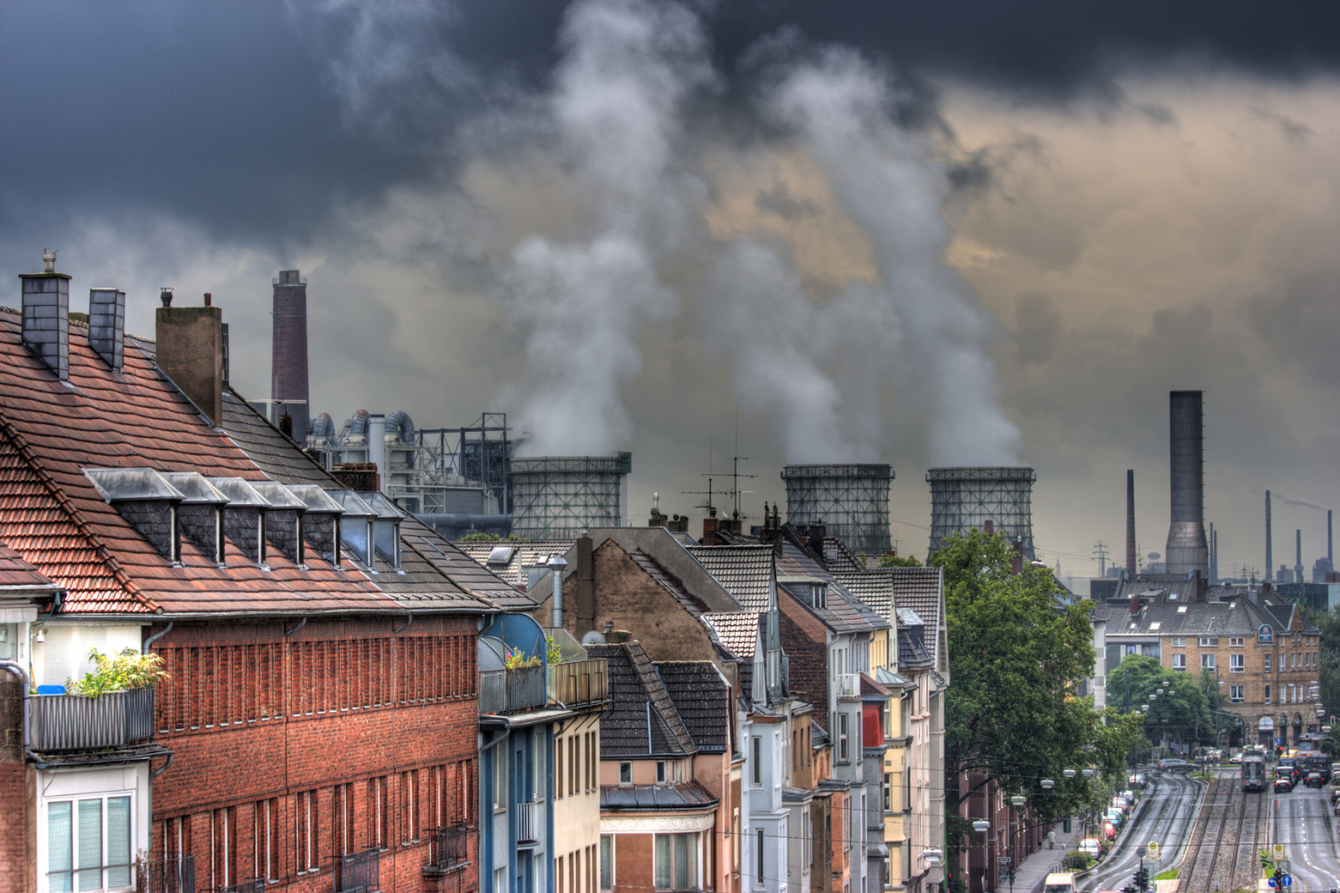 Schöne Bausubstanz im Vordergrund, davor eine Allee mit Straßenbahnschienen, im Hintergrund rauchende Industrieschlote; Detektive der Kurtz Detektei Düsseldorf