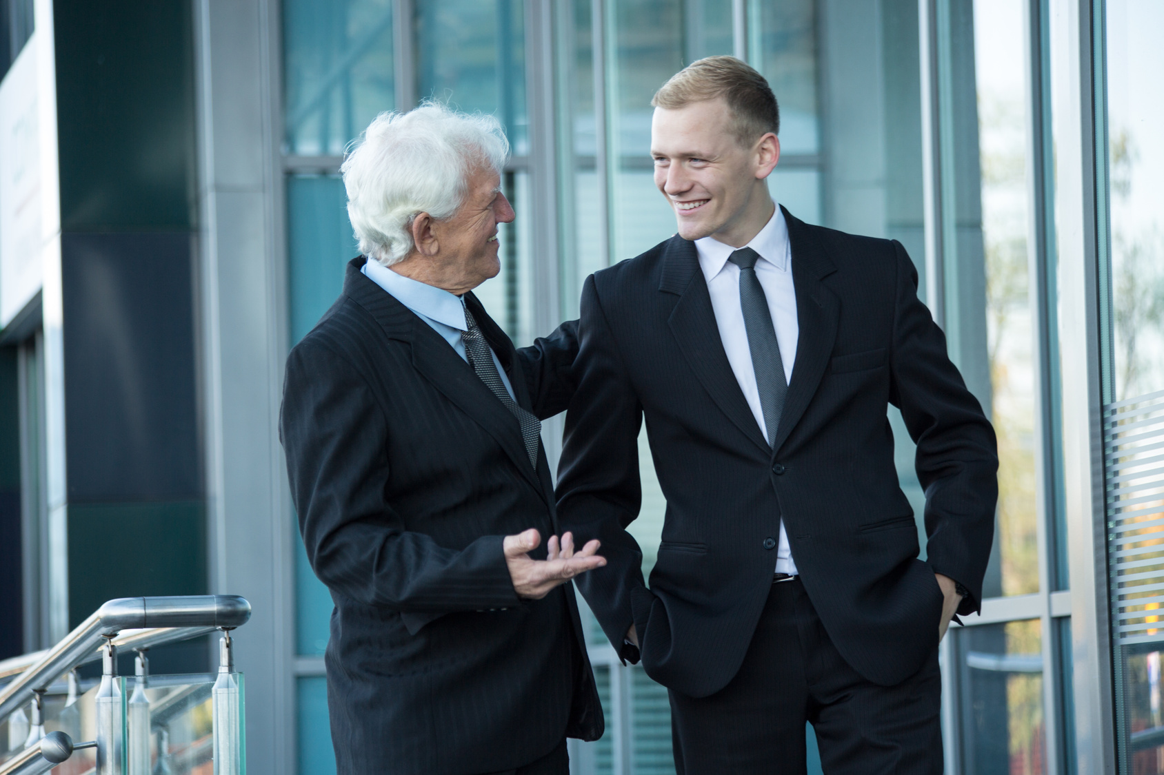 Ein alter und ein junger Geschäftsmann in Anzügen vor einem Bürokomplex, beide lachen; Kurtz Detektei Düsseldorf, Detektiv Düsseldorf, Wirtschaftsermittler in Düsseldorf