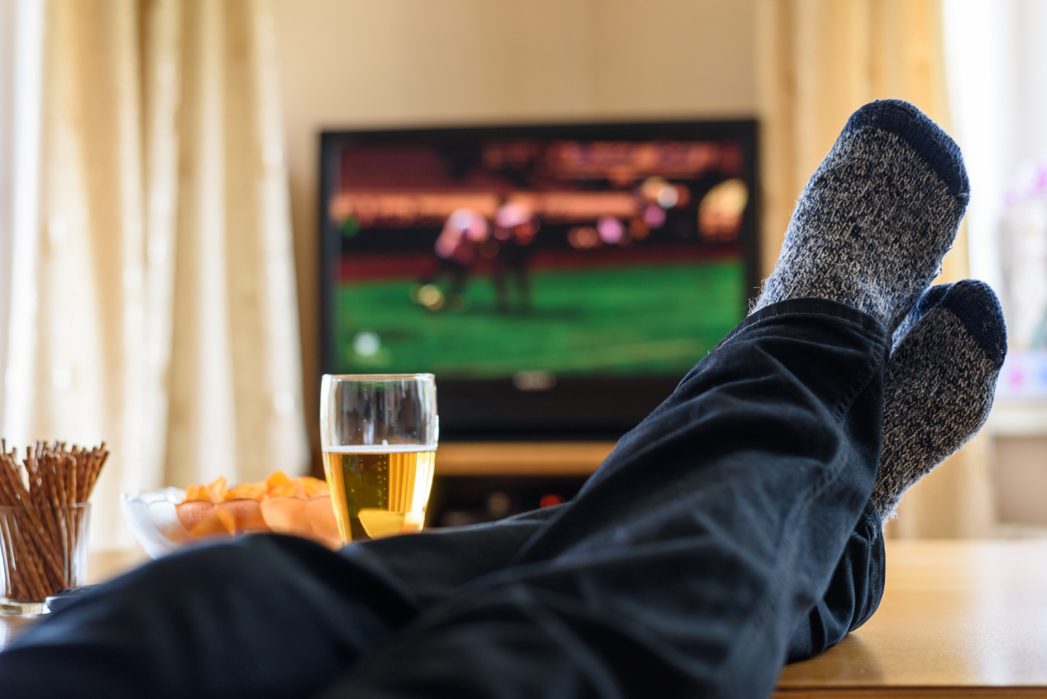 Männerfüße in Strümpfen auf einem Couchtisch, darüber eine lockere-Jeanshose, im Hintergrund läuft Fußball auf einem Fernseher; Bier, Chips und Salzstangen stehen auf dem Tisch; Kurtz Wirtschaftsdetektei Düsseldorf, Detektiv Düsseldorf, Detektivagentur Düsseldorf